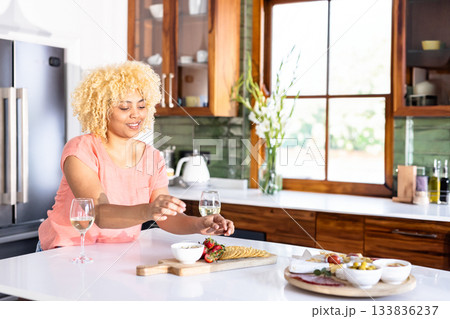 Woman in pink top arranging charcuterie on cutting board beside wine glasses in modern kitchen Woman in pink top arranging charcuterie on cutting board beside wine glasses in modern kitchen 133836237