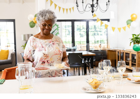 African American senior woman holding platter with meats and cheese among balloons at home African American senior woman holding platter with meats and cheese among balloons at home 133836243