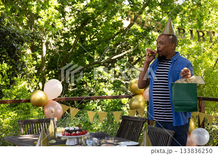 African American man standing on wooden deck holding green gift bag blowing party horn, copy space African American man standing on wooden deck holding green gift bag blowing party horn, copy space 133836250