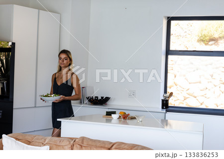 Female holding salad bowl while standing inside home kitchen by island and refrigerator, copy space Female holding salad bowl while standing inside home kitchen by island and refrigerator, copy space 133836253