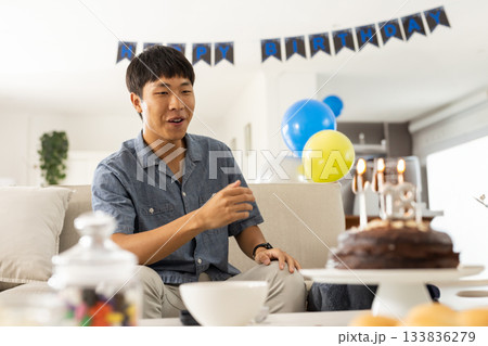 Asian man sitting on sofa marking birthday with cake, balloons, birthday banner, copy space 133836279