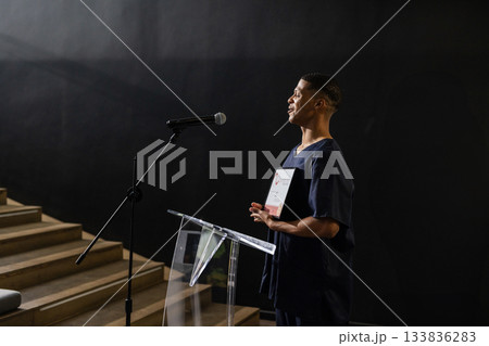 Man standing beside acrylic lectern holding certificate, using mic on stage, copy space Man standing beside acrylic lectern holding certificate, using mic on stage, copy space 133836283