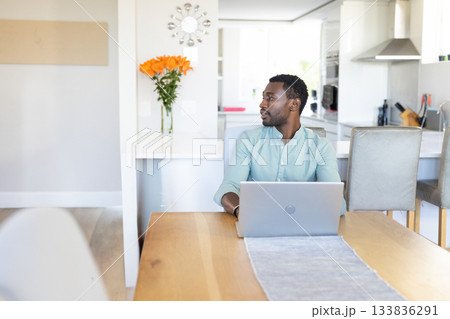 African American man working on laptop at home kitchen dining table with orange flowers, copy space 133836291