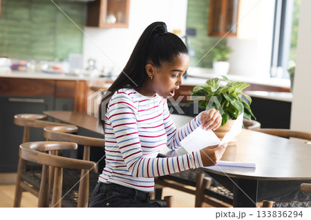 African American woman reading letter at dining table in kitchen beside notebook and plant African American woman reading letter at dining table in kitchen beside notebook and plant 133836294