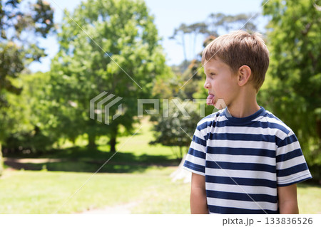 Boy standing on right side in sunny park wearing blue-white shirt, sticking tongue out, copy space Boy standing on right side in sunny park wearing blue-white shirt, sticking tongue out, copy space 133836526