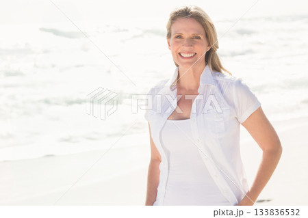 Senior woman smiling while standing on sandy shore in white shirt under bright sunlight, copy space Senior woman smiling while standing on sandy shore in white shirt under bright sunlight, copy space 133836532