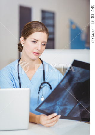 Female doctor wearing scrubs and stethoscope, holding X-ray film, checking laptop at clinic desk Female doctor wearing scrubs and stethoscope, holding X-ray film, checking laptop at clinic desk 133836549