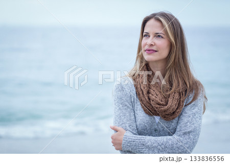Woman standing on sandy beach wearing gray knit cardigan, brown scarf, gazing at sea, copy space 133836556