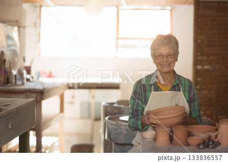 Senior woman wearing apron holding clay bowl at pottery wheel smiling in studio, copy space 133836557