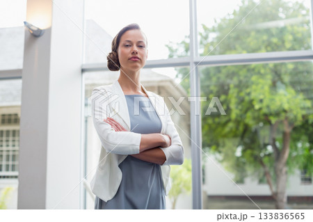 Professional woman standing with arms crossed by wall light in office lobby showing glass windows Professional woman standing with arms crossed by wall light in office lobby showing glass windows 133836565