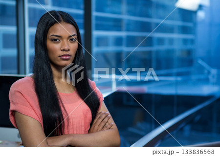 African American woman wearing coral blouse standing with arms crossed in office beside monitor 133836568