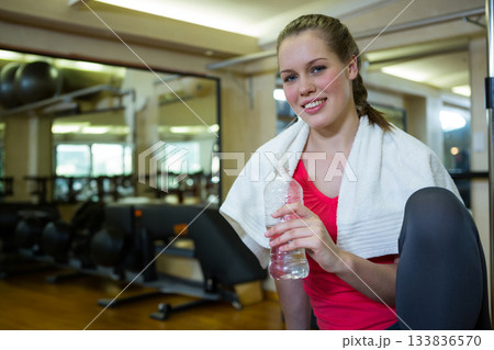 Female exerciser kneeling at gym holding water bottle and wearing towel by wall mirrors, copy space 133836570