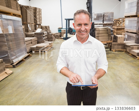 Middle-aged supervisor in workwear checking tablet in warehouse with forklift and cardboard stacks 133836603
