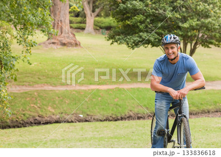 Man leaning on black bicycle wearing multi-color helmet near dirt walking path in park, copy space 133836618
