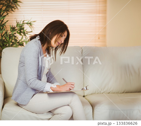 Female consultant wearing business attire writing on clipboard on couch in office, copy space Female consultant wearing business attire writing on clipboard on couch in office, copy space 133836626