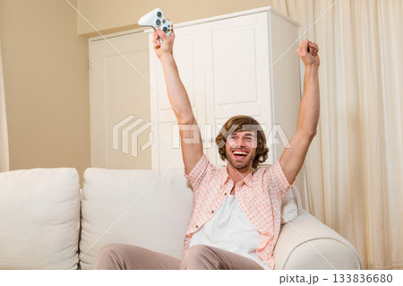 Adult man raising arms triumphantly sitting on white sofa in living room with white game controller 133836680