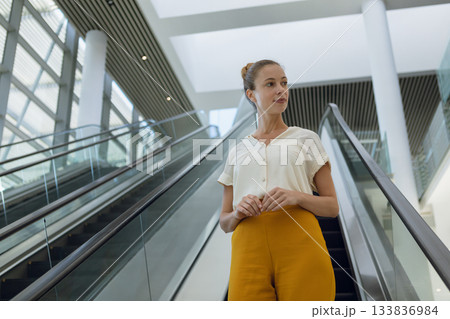 Woman standing on descending escalator in office lobby wearing blouse, yellow pants under skylight Woman standing on descending escalator in office lobby wearing blouse, yellow pants under skylight 133836984
