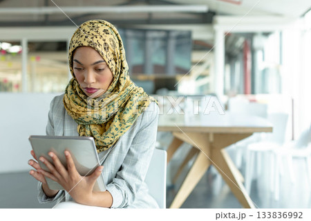 Middle Eastern woman wearing hijab and blazer using tablet at table in coworking office, copy space 133836992