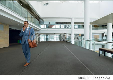 Businessman in suit walking in atrium using handheld device and carrying messenger bag, copy space Businessman in suit walking in atrium using handheld device and carrying messenger bag, copy space 133836998