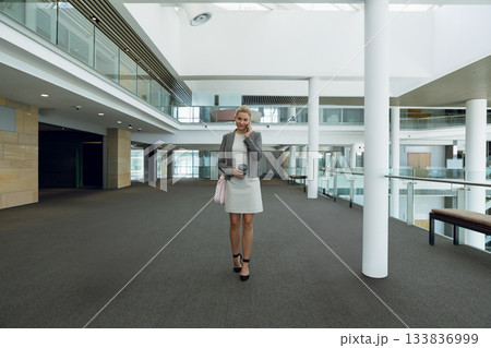 Businesswoman in blazer talking on smartphone, holding coffee cup and carrying pink bag in atrium Businesswoman in blazer talking on smartphone, holding coffee cup and carrying pink bag in atrium 133836999