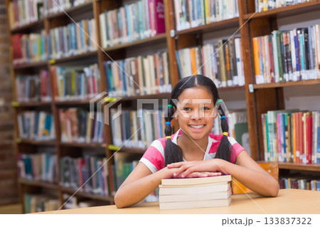 Child female wearing T-shirt sitting at library table among bookshelves resting arms on book stack Child female wearing T-shirt sitting at library table among bookshelves resting arms on book stack 133837322
