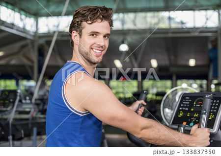Man smiling and exercising on elliptical trainer with digital display under overhead lights in gym Man smiling and exercising on elliptical trainer with digital display under overhead lights in gym 133837350