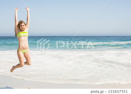 Woman jumping on white sand wearing yellow-green striped bikini with ocean waves behind, copy space Woman jumping on white sand wearing yellow-green striped bikini with ocean waves behind, copy space 133837351