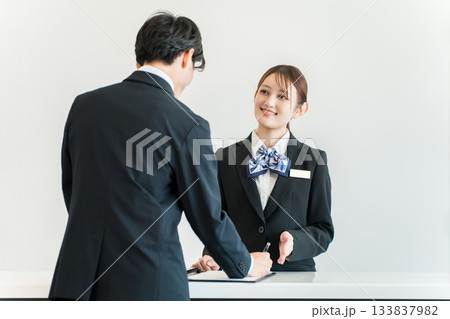 カウンターで手続き,チェックイン,サインする男性と接客対応するホテリエの女性スタッフ カウンターで手続き,チェックイン,サインする男性と接客対応するホテリエの女性スタッフ 133837982