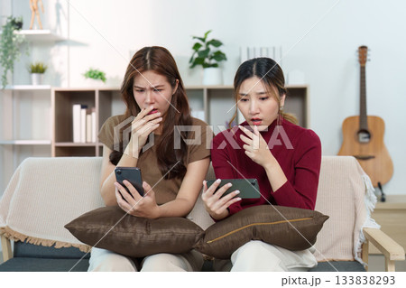 Surprised Reactions. Two friends looking shocked while checking their phones in a cozy living room. Surprised Reactions. Two friends looking shocked while checking their phones in a cozy living room. 133838293