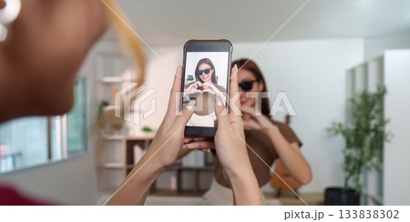 Creative Capture. Young woman posing for a smartphone photo while making a heart gesture. Creative Capture. Young woman posing for a smartphone photo while making a heart gesture. 133838302