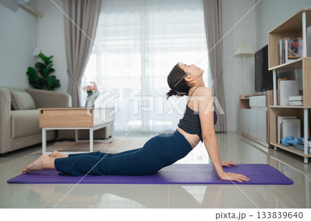 Young woman practicing yoga at home in living room, performing Cobra pose on mat, natural light streaming through windows, peaceful atmosphere 133839640
