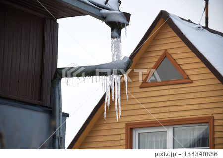 Icicles Hanging From Snowy Roof Pipes Between Dark Wooden Building And Bright Wooden Cottage Icicles Hanging From Snowy Roof Pipes Between Dark Wooden Building And Bright Wooden Cottage 133840886