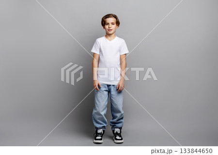 Full-length portrait of young boy standing in studio, wearing white t-shirt, trendy sneakers and jeans, looking at camera with relaxed expression on clean grey background Full-length portrait of young boy standing in studio, wearing white t-shirt, trendy sneakers and jeans, looking at camera with relaxed expression on clean grey background 133844650