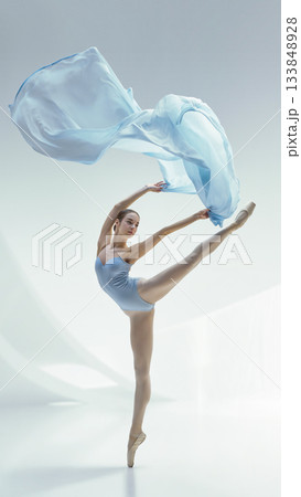 Ballet dancer in high arabesque with flowing blue fabric on minimal light background 133848928