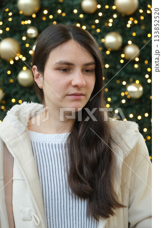 Thoughtful Young Woman in Cozy Coat Against Sparkling Christmas Tree Background. Thoughtful Young Woman in Cozy Coat Against Sparkling Christmas Tree Background. 133852520