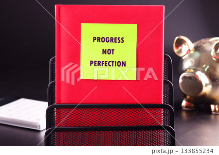 In a sleek workspace, a vibrant red notebook is prominently displayed with a note reading Progress Not Perfection. A golden piggy bank adds a playful touch, inspiring growth and change In a sleek workspace, a vibrant red notebook is prominently displayed with a note reading Progress Not Perfection. A golden piggy bank adds a playful touch, inspiring growth and change 133855234