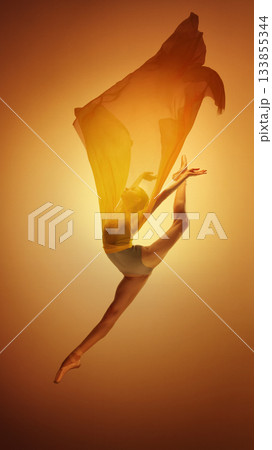 Ballet dancer performing high leap with golden fabric on warm orange backdrop Ballet dancer performing high leap with golden fabric on warm orange backdrop 133855344
