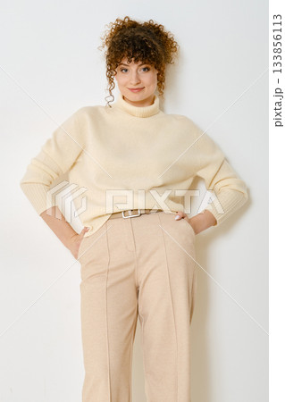 Stylish woman in cream sweater and beige pants poses confidently against a white background 133856113