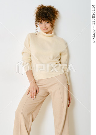 Stylish woman poses in beige sweater and trousers against a plain white background 133856114