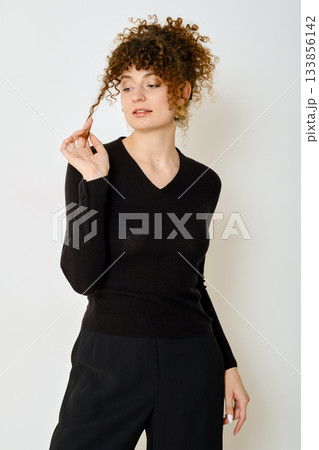 Woman with curly hair in a black sweater poses thoughtfully against a plain background 133856142