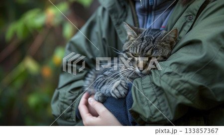 A young person with short hair holds a small tabby cat in their arms. The background features blurred greenery, suggesting an outdoor setting. 133857367