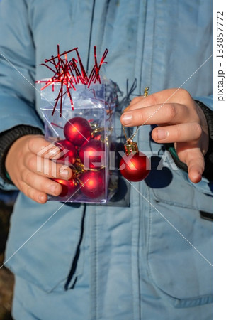 Close-up of hands holding a box with Christmas tree ornaments and a single round bauble in the other hand. Festive holiday preparation, winter decoration, and family seasonal activities. Close-up of hands holding a box with Christmas tree ornaments and a single round bauble in the other hand. Festive holiday preparation, winter decoration, and family seasonal activities. 133857772