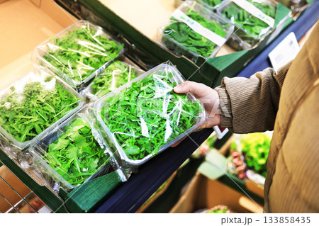 A person's hand selects a clear plastic container of fresh green arugula leaves from a refrigerated shelf in a grocery store. High quality photo 133858435