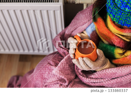 A close-up of a girl holding a cup of tea. She sits near a radiator. She's wearing a scarf, gloves, and a sweater. She's wrapped in a purple blanket. She's trying to keep warm in a poorly heated room, 133860337