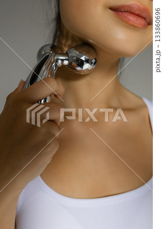Close-up of a young woman massaging her neck with a metal 3D roller, demonstrating a tightening and relaxing skincare routine at home. Close-up of a young woman massaging her neck with a metal 3D roller, demonstrating a tightening and relaxing skincare routine at home. 133860696