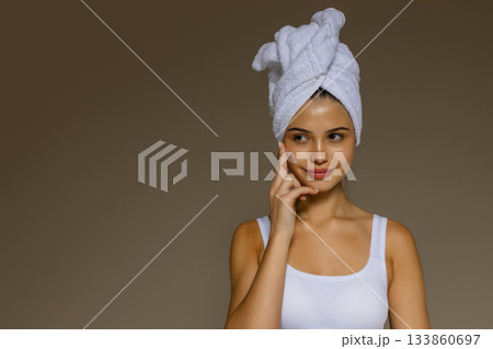Young woman applying skincare cream to her cheek after a shower, wearing a towel on her head and showing a gentle beauty routine. Young woman applying skincare cream to her cheek after a shower, wearing a towel on her head and showing a gentle beauty routine. 133860697