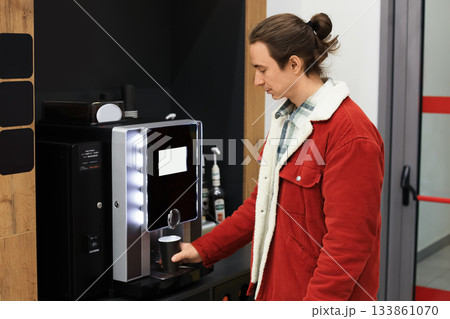 Young man in a red jacket is using an automatic coffee machine in an office or waiting area for a hot drink. High quality photo 133861070