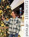 Young man in plaid shirt taking a selfie next to a large, brightly decorated Christmas tree in a modern shopping mall. High quality photo 133861072