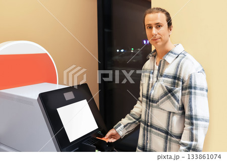 A young man in a plaid shirt uses a self-service terminal, inserting a card, possibly at an airport or station. High quality photo A young man in a plaid shirt uses a self-service terminal, inserting a card, possibly at an airport or station. High quality photo 133861074