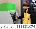 Young man in a bright yellow winter jacket and hat operating an ATM touchscreen, withdrawing or depositing money. High quality photo 133861076